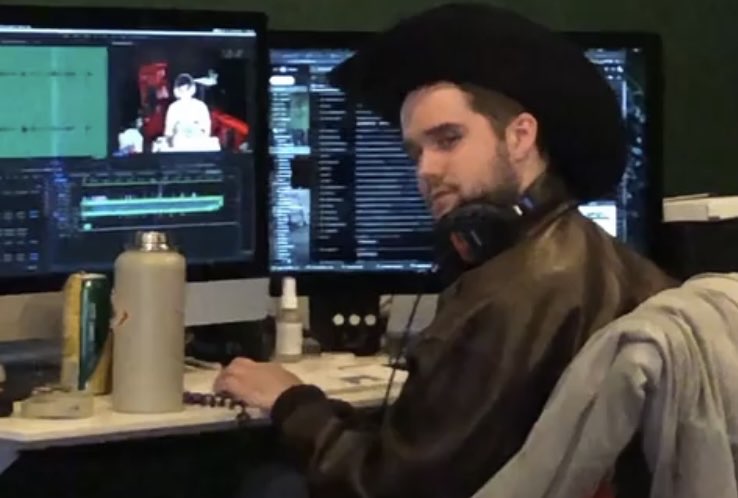 spencer looking over his shoulder with an exaggerated smouldering expression while he edits. he wears a cowboy hat, headphones around his neck, and a leather jacket while seated at a desk with two monitors showing editing software.